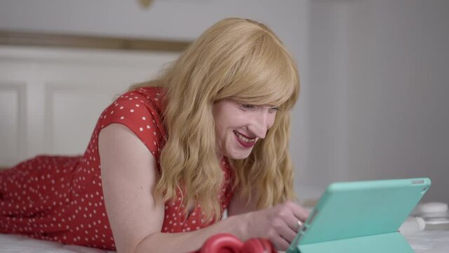 Portrait of relaxed trans woman lying on bed messaging online on tablet smiling. Happy Caucasian LGBTQ transgender person in red dotted dress surfing Internet at home in bedroom. Individuality and joy