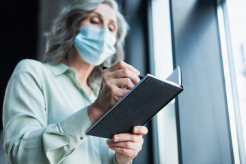 Fototapeta na wymiar Low angle view of blurred businesswoman in medical mask writing on notebook in office.