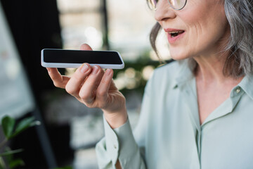 Cropped view of grey haired businesswoman recording voice message on cellphone in office.