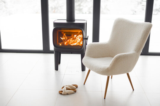 Cozy Living Space By The Burning Fireplace With Chair, Cup And Slippers On Background Of Snowy Landscape. Idea Of Home Comfort And Winter Mood