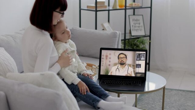 Telemedicine Chat. Mom And Her Sick 4 Years Old Girl Talking On Conference Video Call To Male Caucasian Doctor. Virtual Therapist Consulting Family During Online Appointment On Laptop At Home.