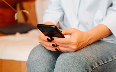 Close-up of female hands holding black mobile phone, indoors. Caucasian woman chatting, typing, browsing internet on smartphone while sitting on couch at home. Selective soft focus