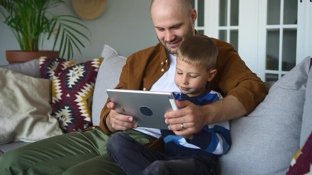 Father and toddler online development education. Young bearded man spbd shows educative video to cute toddler via tablet sitting on sofa in living room closeup