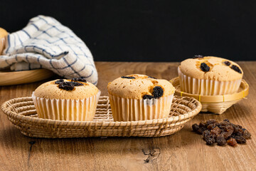 Homemade raisin sponge cupcake in bamboo basket with pile of dried raisin, wooden tray and table cloth on wooden table on black background.