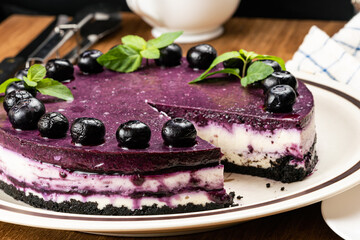 Closeup part of delicious homemade blueberry cheesecake garnished with preserved blueberry and green mint leaf in brown ceramic dish with metal spoon, white ceramic coffee cup on wooden table.