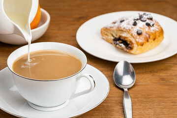 Pouring fresh milk into coffee in white ceramic cup on ceramic saucer with Danish Pastry filled with chocolate cream topping with chocolate chips and icing sugar in ceramic dish and ripe red apple.