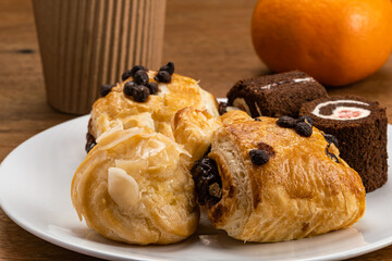Various baked pastry dessert, Eclair filled with corn cream, Danish Pastry filled with sweet chocolate cream and Black Forest Chocolate Cake Roll in white ceramic dish with ripe orange and coffee cup.