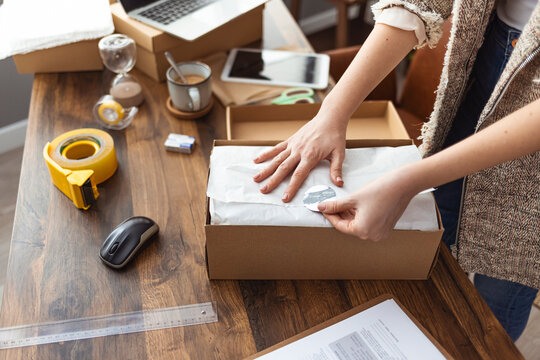 Working Woman At Online Shop. She Wearing Casual Clothing And Checking On Laptop Address Of Customer And Package Information. Young Woman Selling Products Online And Packaging Goods For Shipping.