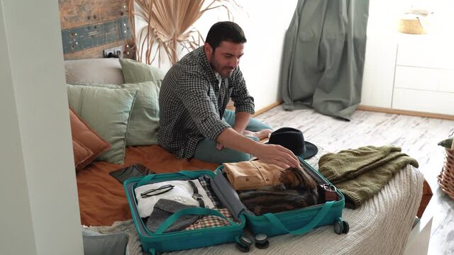 Happy man packs winter themed cyan bag for holiday and petting cat. Vacation preparation, careful bag packing. Millennial generation Y travel blogger pack suitcase. loft design room. 4k footage