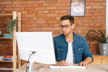 Portrait of focused young man wearing stylish glasses working on desktop computer sitting at desk...