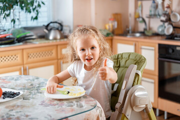 Happy little girl child show gesture of approval, Thumbs up by finger after eating food in kitchen...