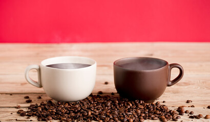 Two brown and off white coffee cups with smoke and beans on rustic wooden table, red background.