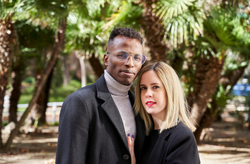 Black man and blond woman in outerwear looking at camera while standing against palms during romantic date on sunny day in exotic park