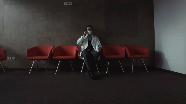 A young doctor in a white work coat and respirator sits in a dark room and consults over the phone