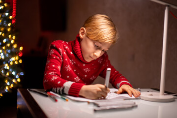 a boy in a red Christmas sweater on the background of a Christmas tree diligently draws behind a lamp that shines on him