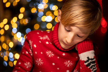 a boy in a red Christmas sweater on the background of a Christmas tree pressed his hand to his face and thoughtfully draws