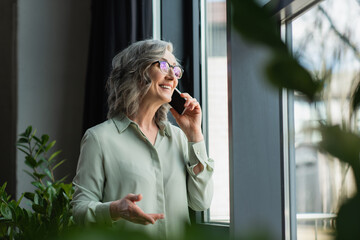 Positive businesswoman talking on smartphone near plants in office.
