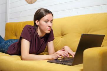 A young girl lies at home on the couch and works on a laptop. Homeschooling, remote work, freelancer