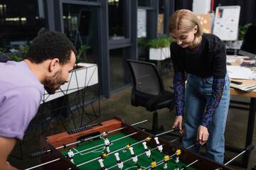 blurred african american manager playing table football with happy blonde colleague.