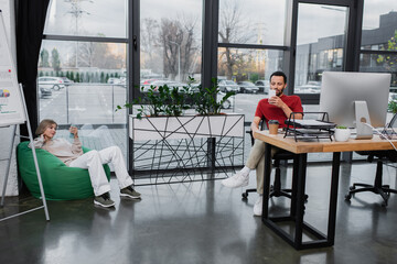 Fototapeta premium blonde woman sitting in bean bag chair and using smartphone near african american colleague in office.