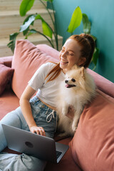 Vertical shot of cheerful young woman using laptop sitting on comfortable sofa cuddling cute small...