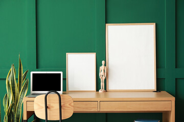 Modern workplace with laptop, mannequin and blank frames near green wall