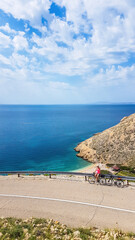 A girl in a sporty outfit, standing on the bike, on the side of a steep road. In the bottom Mediterranean Sea. Girl is enjoying the view. Thick clouds above. Active holidays. Hidden gem of Croatia