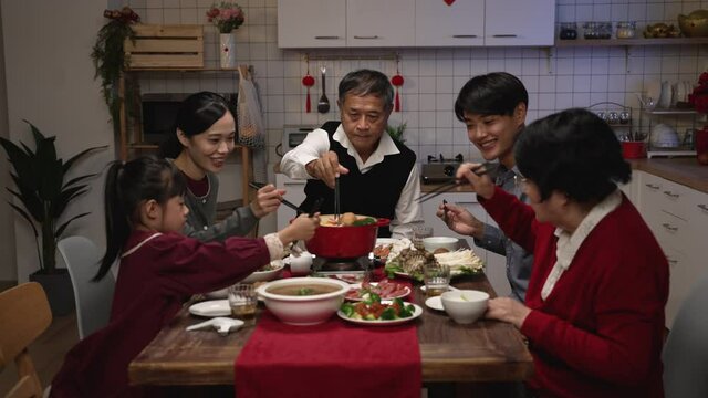 happy extended family putting ingredients with chopsticks while cooking hot pot together during chinese lunar new year reunion dinner at home