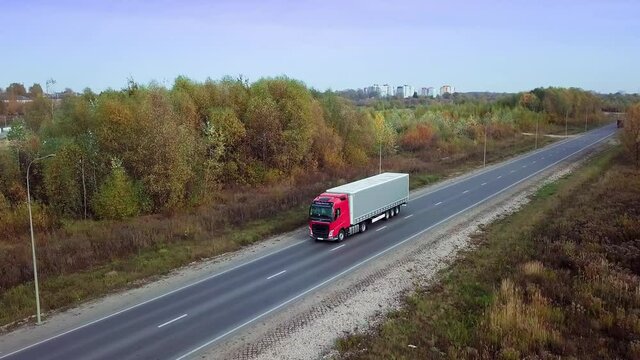 Aerial front side view of Red flat-nose truck with gray cargo trailer pulls onto the highway from the roadside. International cargo delivery. Autumn nature in the sunset light