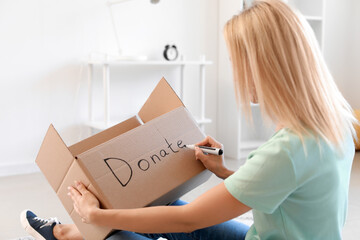 Young woman writing word DONATE on cardboard box at home