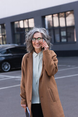 Cheerful mature businesswoman in coat talking on cellphone and holding documents on urban street.