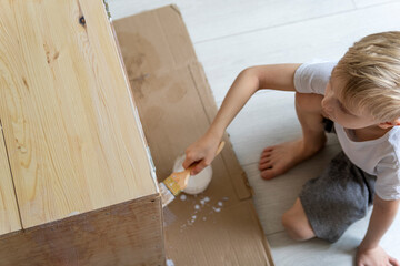 Top view boy blond paints wooden pedestal with brush. Child in carpentry workshop.