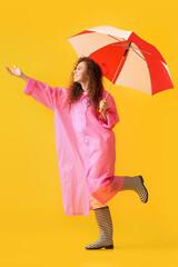 Young African-American woman wearing pink raincoat and gumboots with umbrella on yellow background