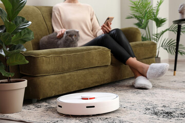 Modern robot vacuum cleaner near sofa with resting woman in room