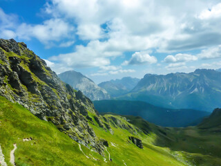 A stunning view on Alpine valley. Hiking trails in the high mountain. Green grass and bushes cover the slopes. Endless chains of mountains visible. Partially cloudy day. Spring in the mountains.