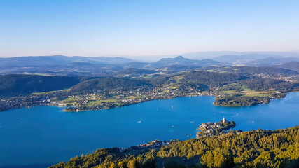 Obraz premium A view on the Woerthersee lake from the observation deck of Pyramidenkogel Tower. Lake is reflecting the last beams of sun for this day. Golden hour. The surrounding hills are shining gold.