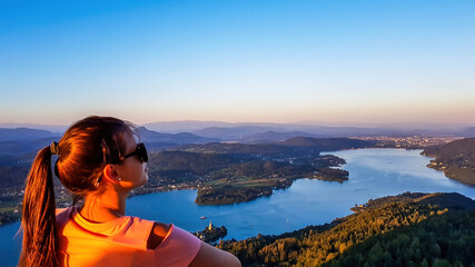 Fototapeta na wymiar A girl standing on an observation deck and enjoying the stunning view on the valley and the Woerthersee lake during the sunset. Hills are catching the last sun beams. Contemplation of the nature