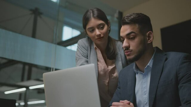 Young middle eastern man newcomer study to work on laptop at new job, young woman helping him at office, tracking shot