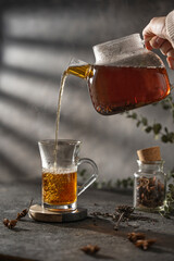 Pouring hot tea into glass cap, beautiful gray textured table with dry spices and  hand holding transparent teapot,  Studio shot with copy space.