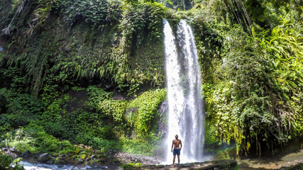 A man in swimsuit standing under two levelled waterfall in Lombok, Indonesia. Tiu Kelep Waterfall...