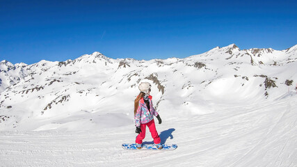 Fototapeta na wymiar A snowboarder going down the slope in Heiligenblut, Austria. Perfectly groomed slopes. High mountains surrounding the girl, wearing pink trousers and colorful jacket. Girl wears helm for protection