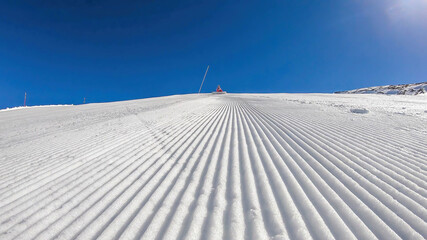 A close up on a perfectly groomed slope, captured on a brisk and bright morning in Heiligenblut, Austria. A girl sitting on top of the slope. Sunbeams reflecting itself on the bright snowy surface.