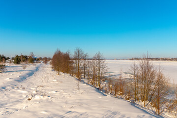 Beautiful winter landscape with river shores, trees, snowy meadow and blue sky in background