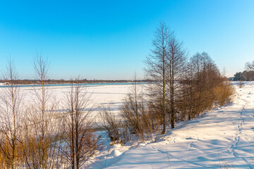 Beautiful winter landscape with river shores, trees, snowy meadow and blue sky in background