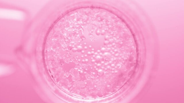 Top view macro shot of crystals fall down from test tube into clear fluid in flask creating different sized bursting bubbles on pink background | Mineral skincare cosmetics formulating concept