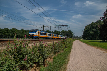 Moving blue yellow white Dutch NS Sprinter Train along a gravel road and the fields of Estate...