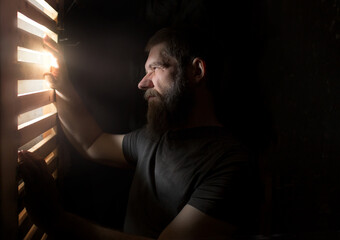 a man with a beard stands in the dark at a door with slits from which a bright light shines on him
