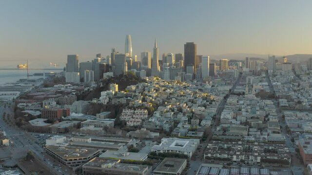 4K aerial video footage of San Francisco, California, at sunset. Movement is retreating towards Pier 39 area. Tops of buildings and houses lit by yellow by setting sun.