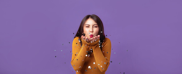 Young Asian woman cheerful blowing confetti on banner isolated purple studio background. Asian female happy in anniversary concept.