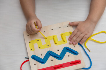 Child plays educational game interestedly with wooden colorful board and laces. Development,...